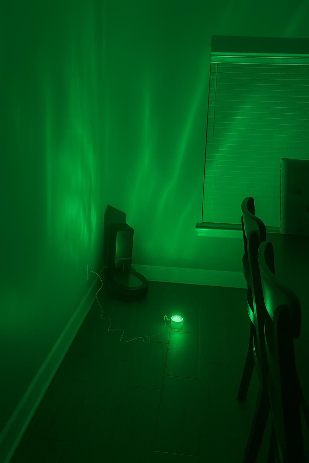 Room with green lighting from an aurora projector lamp on the floor, featuring a window with blinds and a small table.